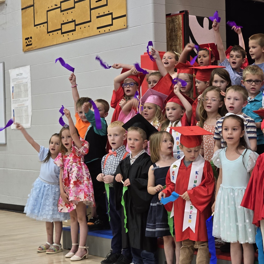 Kids in red hats and colorful dresses stand in a row, raising colorful ribbons. Some wear glasses.
