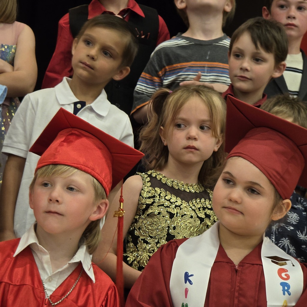 Kids in red graduation caps and gowns stand together. A girl in a gold dress is behind them.