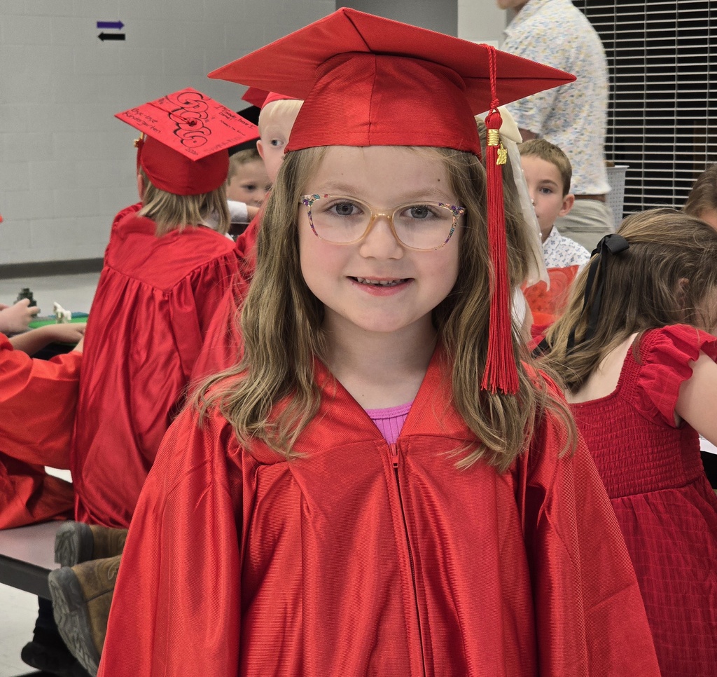 Girl in red cap and gown, smiling at the camera, with other children in red dresses.
