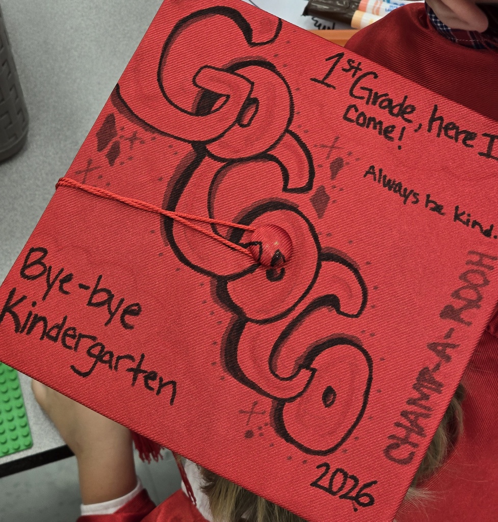 A child holds a red graduation cap that says "Goodbye Kindergarten." The cap is decorated with black letters and numbers.