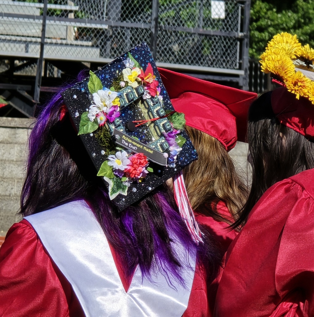Graduates in red caps and gowns stand close together, one with purple hair, another with a floral hat decoration.