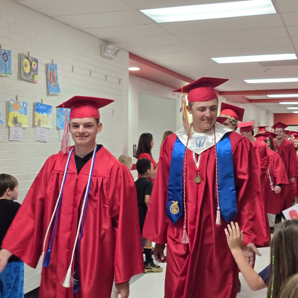 A hallway with red caps and gowns. Two people walk in front. Behind them, others in red caps and gowns walk.