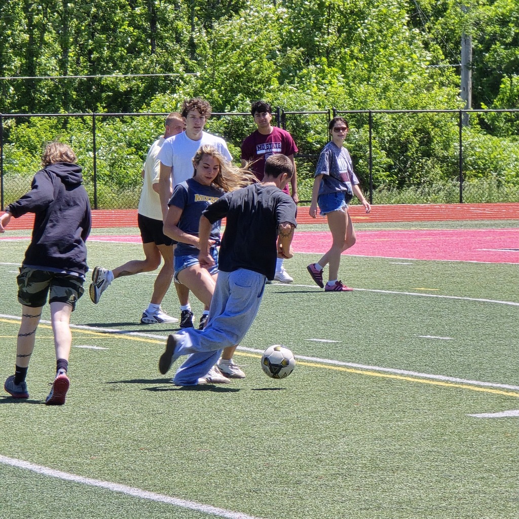 Six individuals play soccer on a field. One person runs toward the ball while others watch.