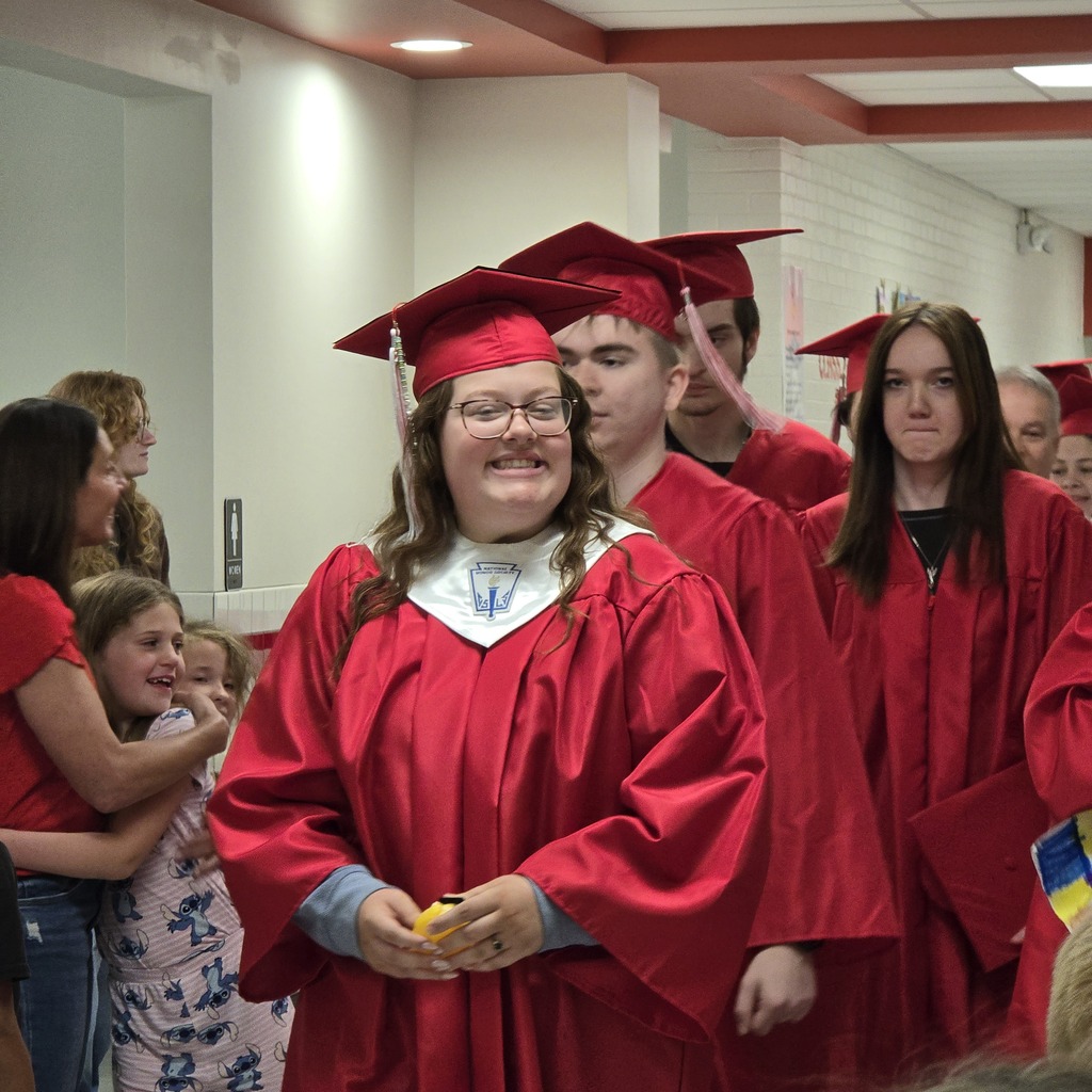 A line of people in red gowns and hats in a hallway. One individual holds a yellow object.