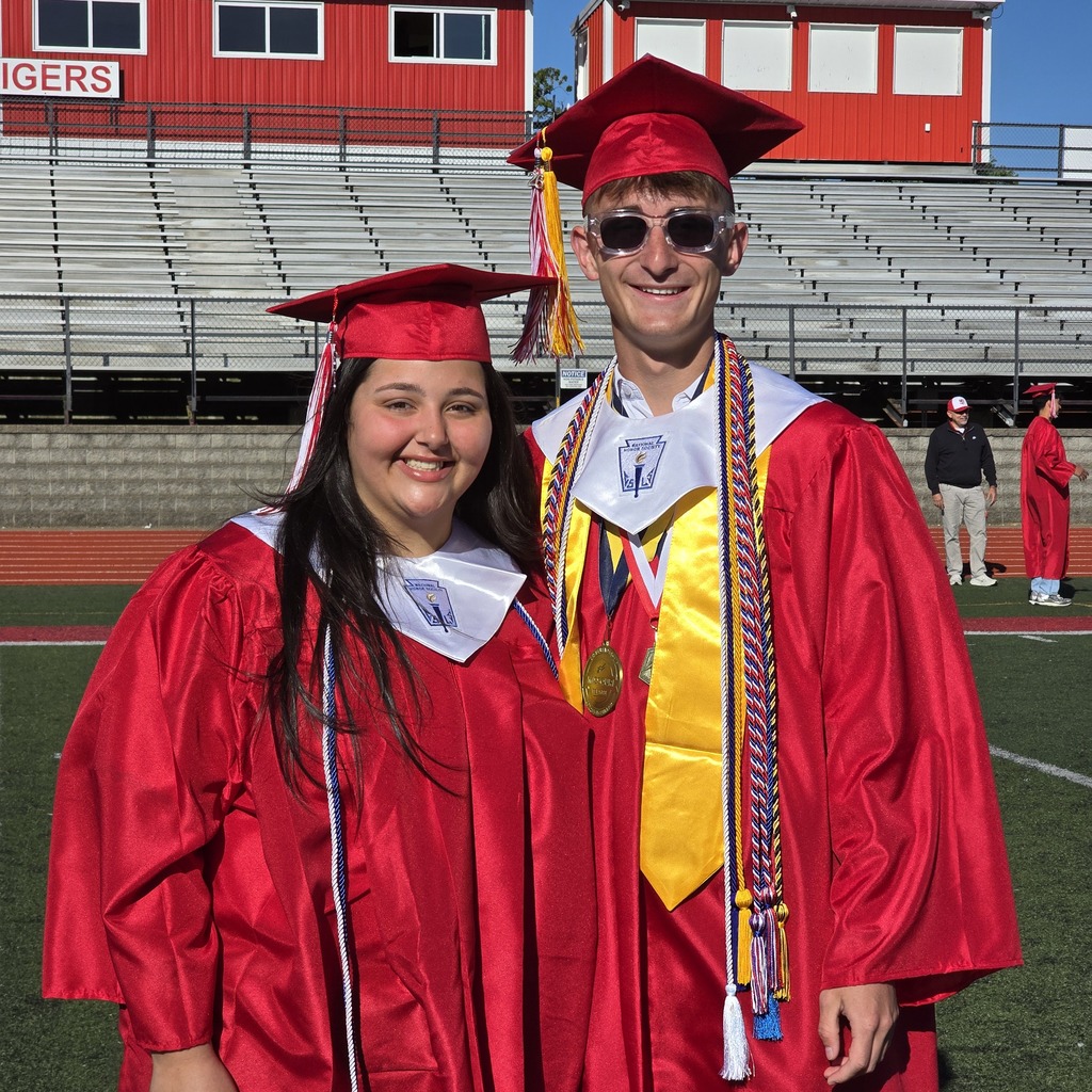 Couple in red gowns and caps stand on a football field. Behind them, a stadium with bleachers.