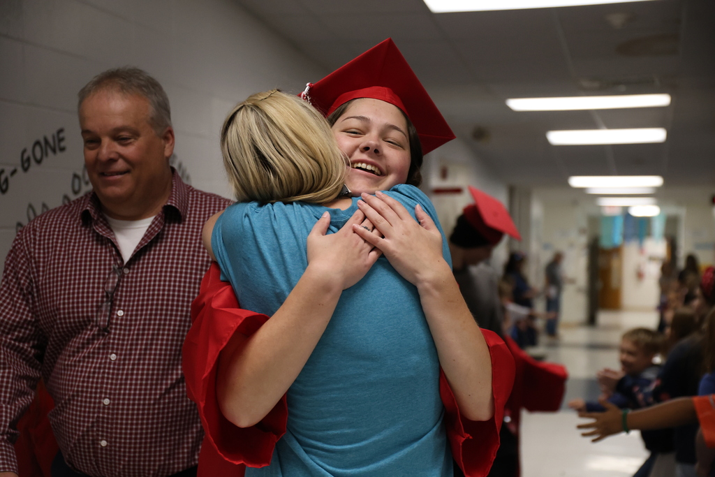 A woman in a blue shirt and red graduation gown hugs another woman in a hallway.