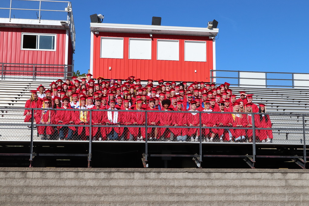 A group of people in red graduation gowns pose for a photo on a stadium bleacher.