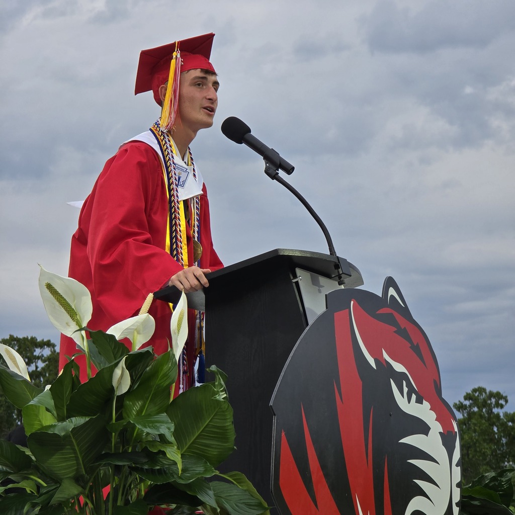 A person in a red gown stands at a podium, speaking into a microphone. A logo of a tiger is behind the podium.