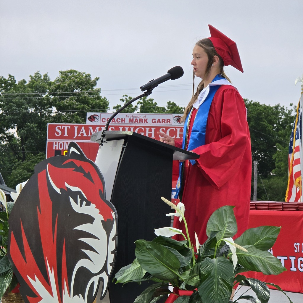 A woman in a red gown speaks into a microphone at a podium, with a red and black tiger emblem.
