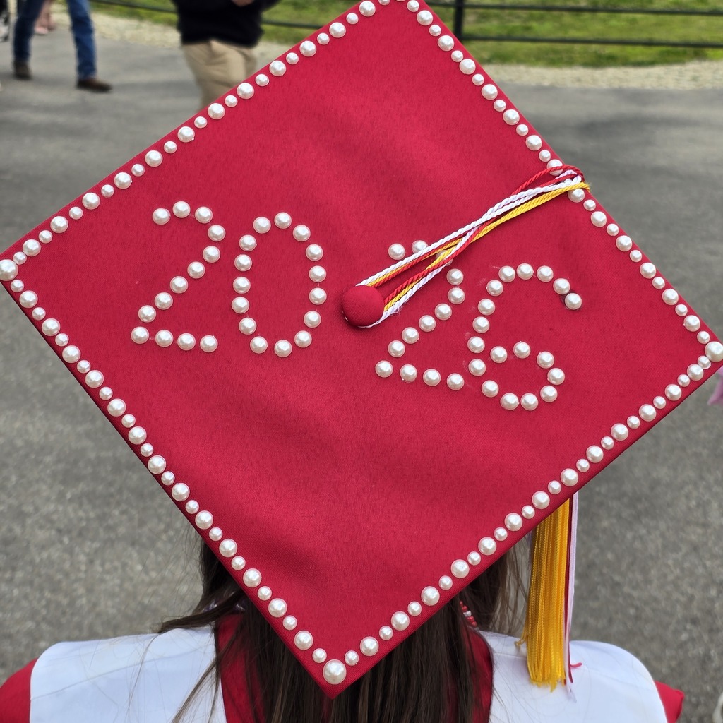 Red cap with white pearls and "2020" in center. Person in white and red, blurry background with people.