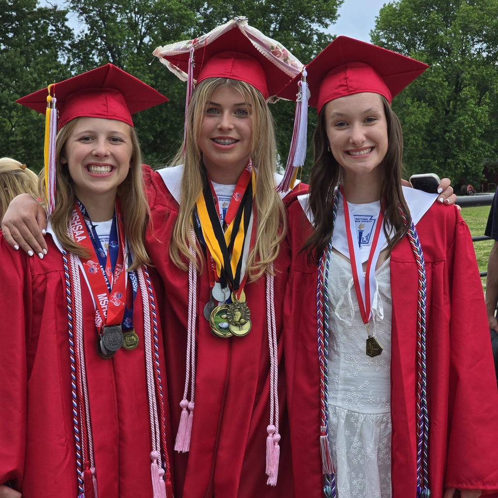 Three women in red graduation gowns and caps pose together outdoors, showcasing their medals.