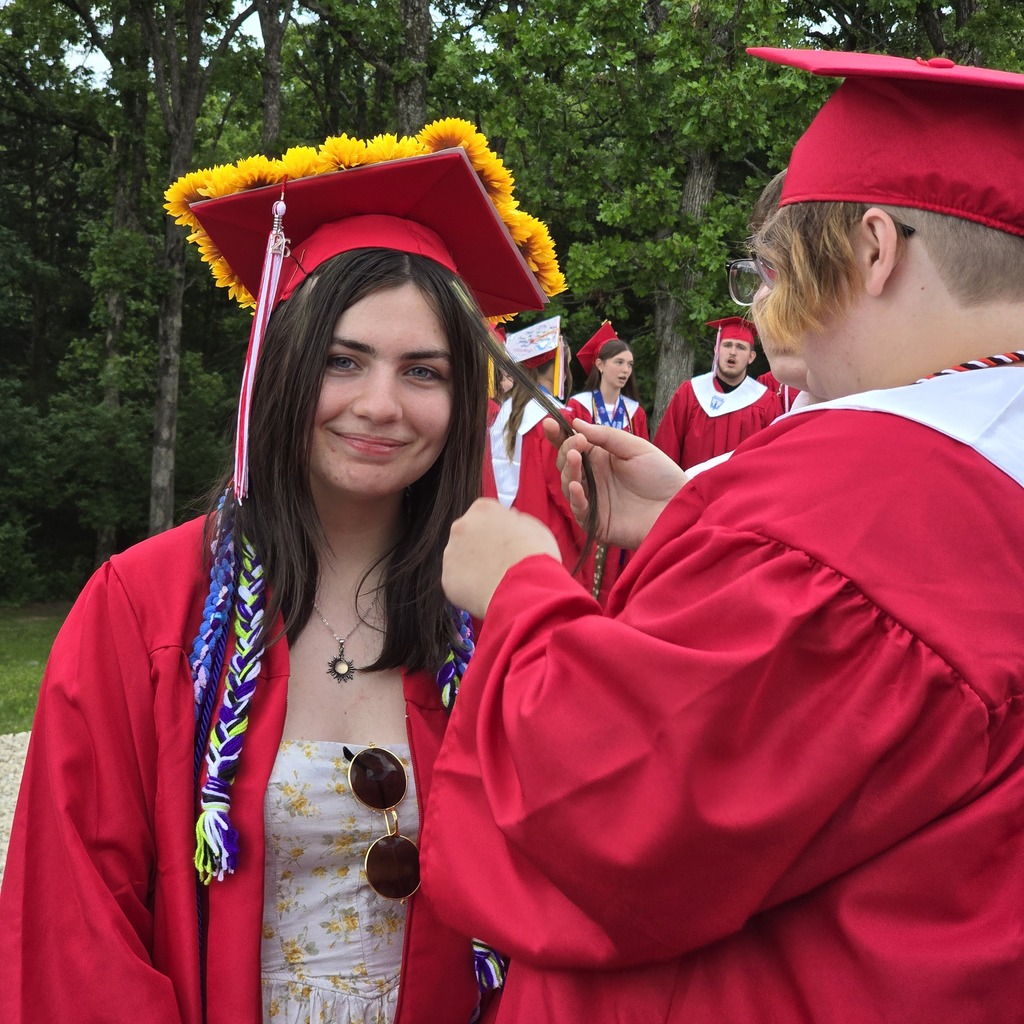 A person in a red graduation gown assists a woman, also in a red gown, with her cap.