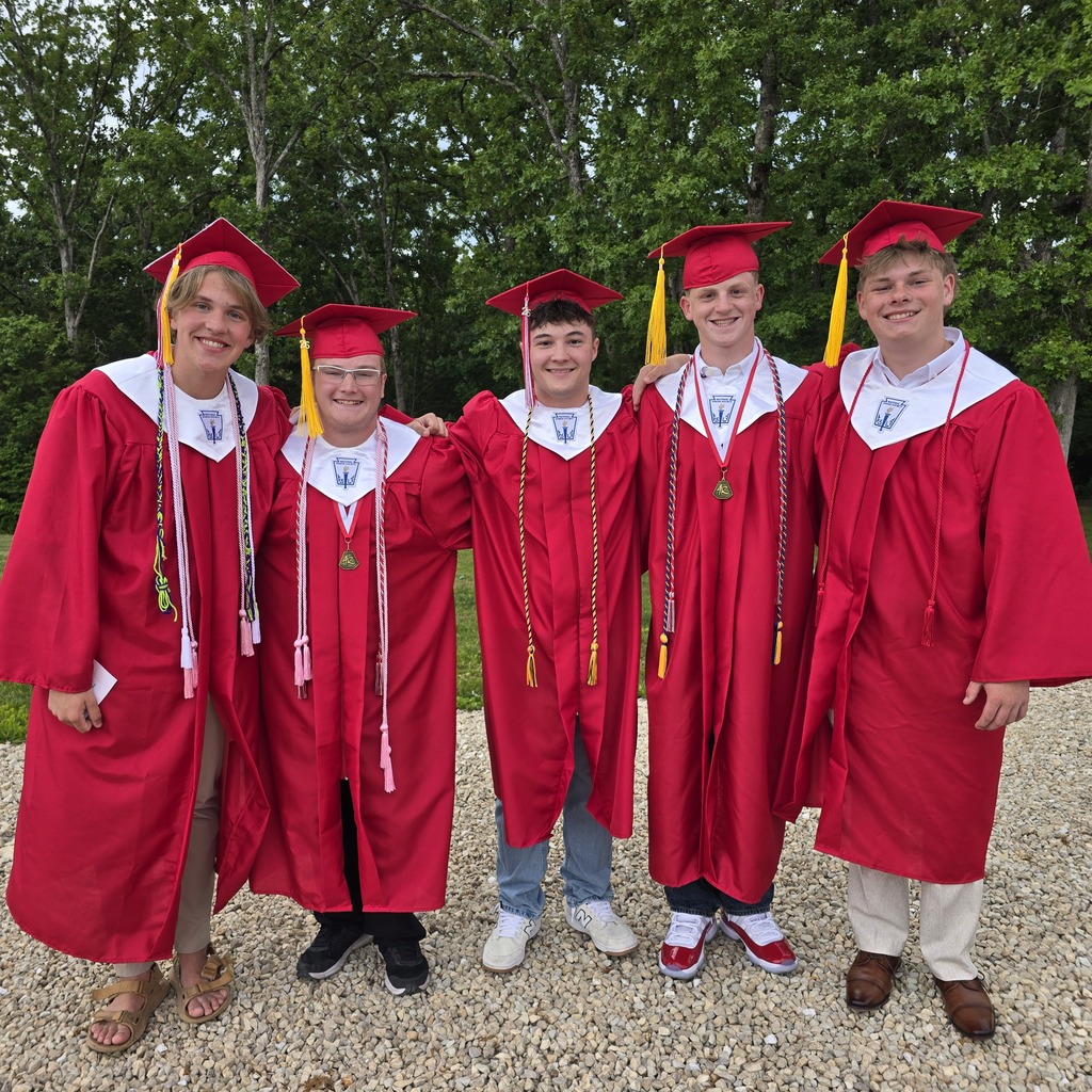Five individuals in red graduation robes and caps stand together on a gravel surface with trees in the background.