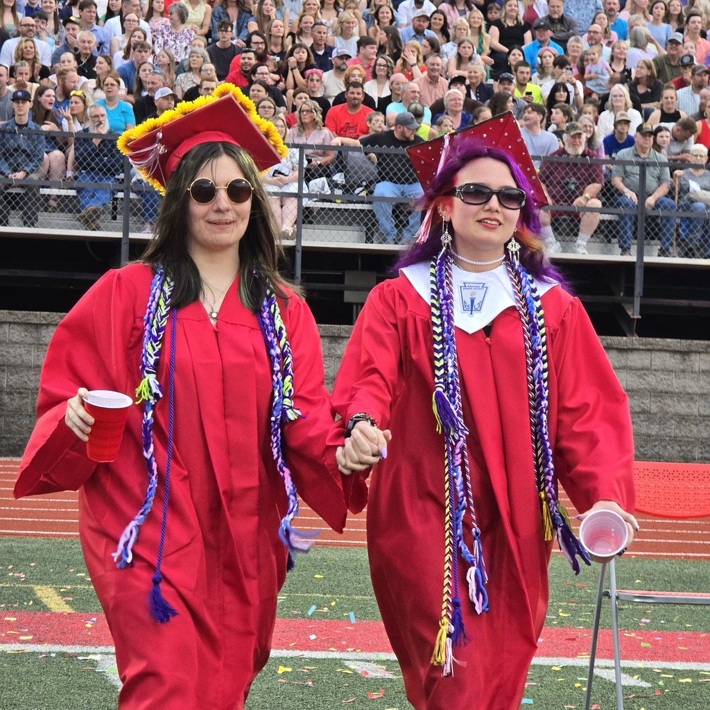 Two women in red gowns and hats walk on a field. They each hold a cup. Behind them, spectators sit on bleachers.