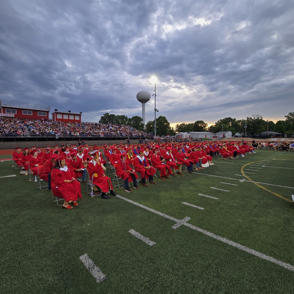 Graduates in red gowns sit in rows on a football field with white lines; cloudy sky above.