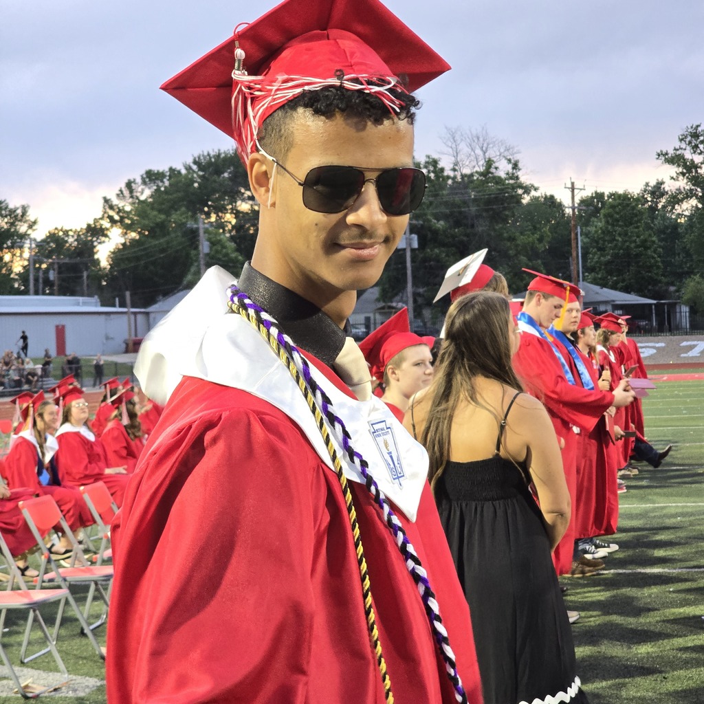 A male graduate in a red cap and gown stands on a field, smiling. Behind him, other graduates wait.
