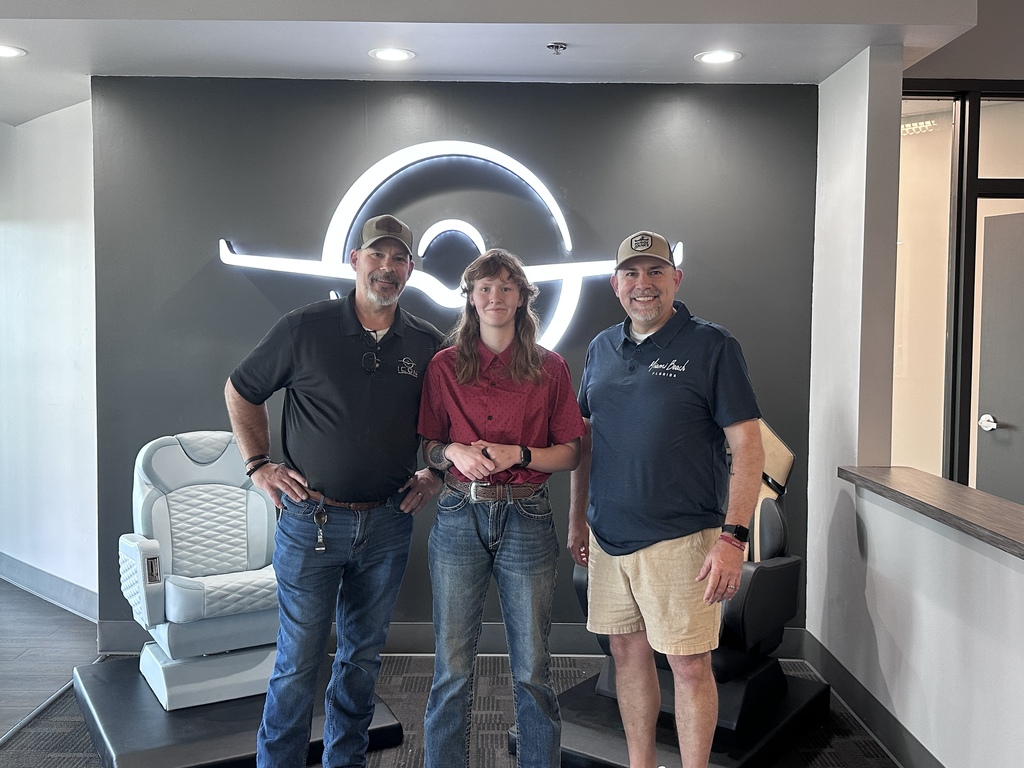 Three people stand in front of a white wall with a large curved neon logo. Two men wear hats and the woman wears a red shirt.