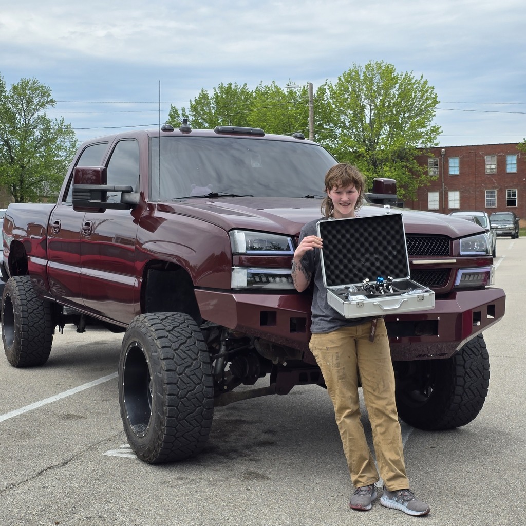 A person holds a case in front of a lifted, maroon truck in an outdoor parking lot.