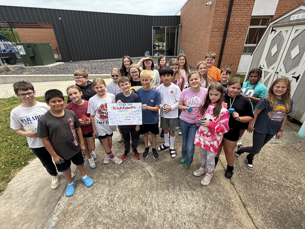 Group of children at a school, standing in a line, holding a white banner. A building with brick walls and a green dumpster.
