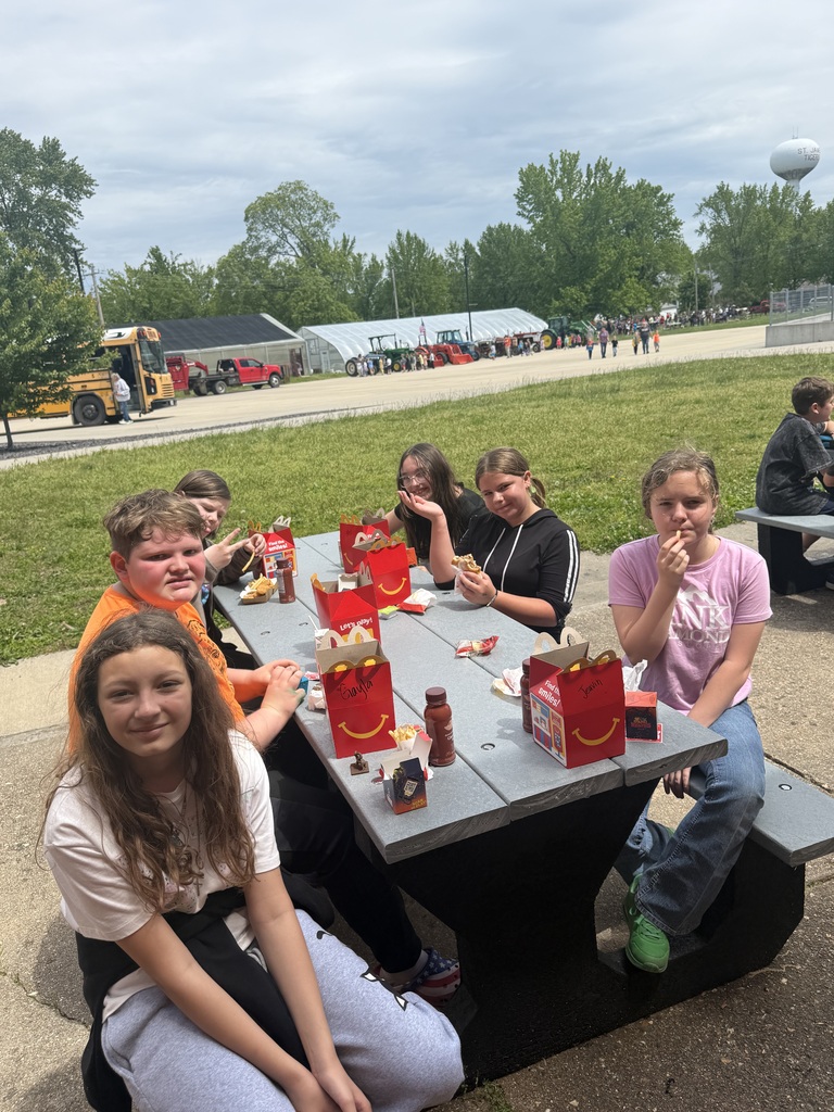 A group of children sitting at a picnic table with red boxes and food, smiling.