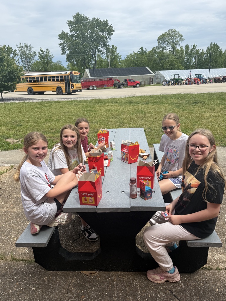 Six girls sitting at a picnic table with food boxes and bottles in front of them. A school bus is parked behind them.