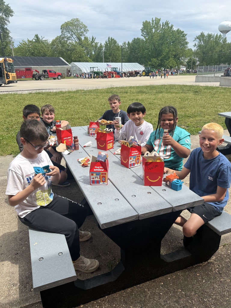Seven children sit at a picnic table, some wearing glasses, with various food items in front of them.