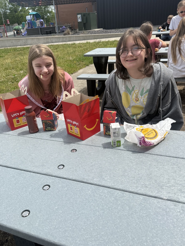 Two girls sit at a table with food in paper bags. They are surrounded by condiments and snacks.