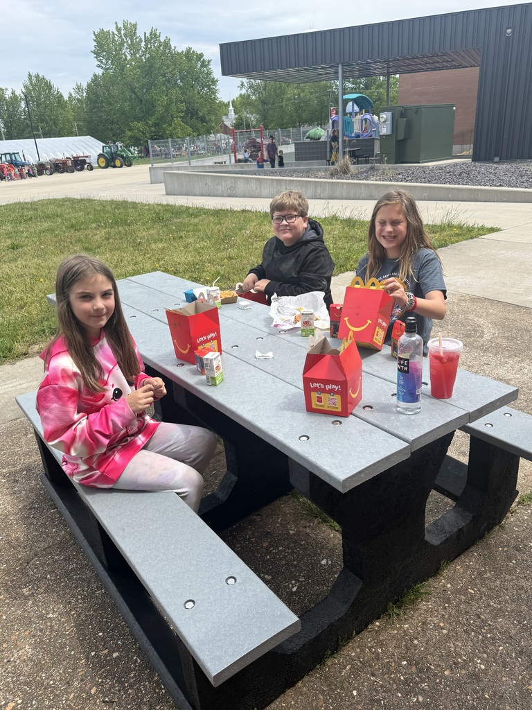 Three children are seated at an outdoor table with food and drinks on it. Behind them is a grassy area with trees.
