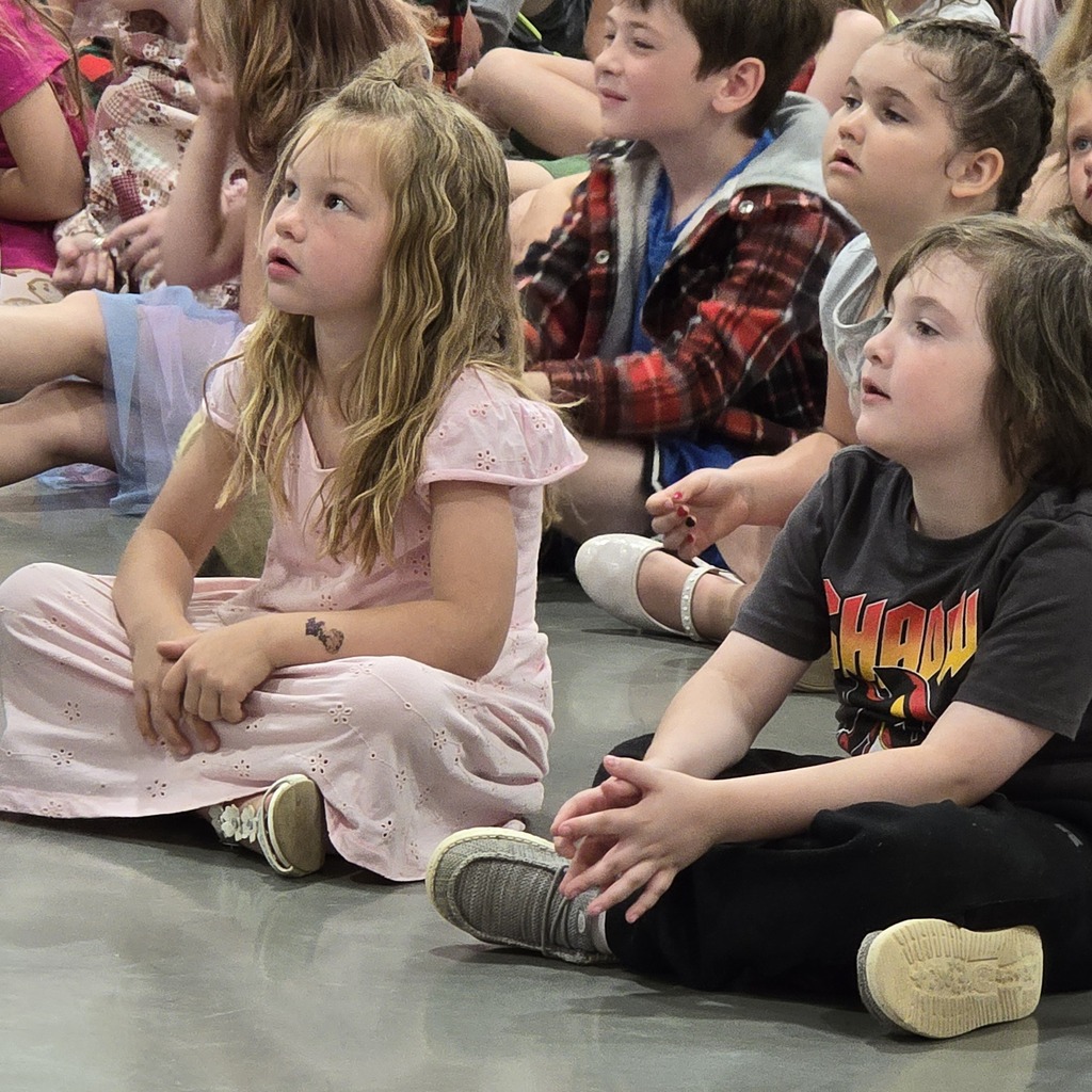 Group of children sitting in a row on a floor, some wearing sneakers and bracelets.