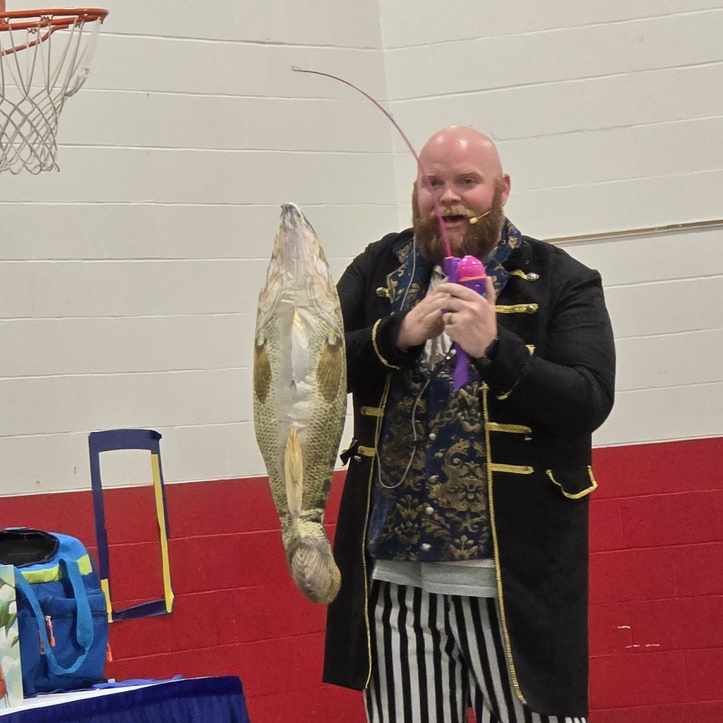 A man dressed in a black coat and striped pants holds a large fish on a stick. He has a purple object in his hand. A basketball hoop is on the wall behind him.