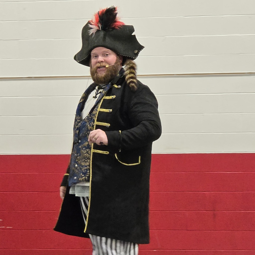 A person in a black coat, white striped pants, and a black hat with a feather stands in front of a red and white wall.