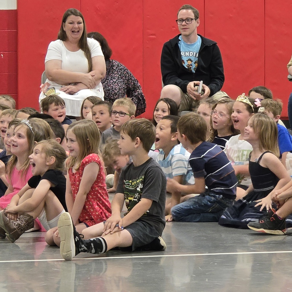 Children and adults sit and stand on a floor, possibly in a school auditorium, with some wearing glasses.