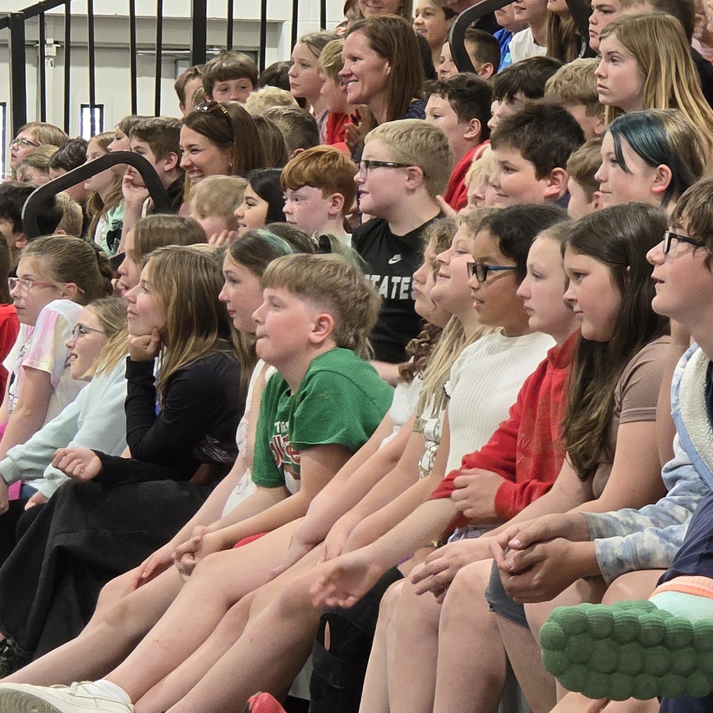 A crowd of children seated in bleachers, with some wearing glasses and sneakers. Some are looking forward, while others appear engaged in conversation.