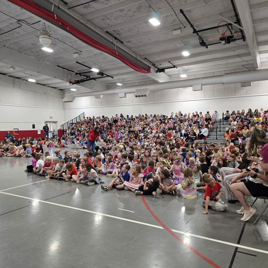 Large indoor space with a crowd of children seated in rows, adults standing, and a person seated on a chair.
