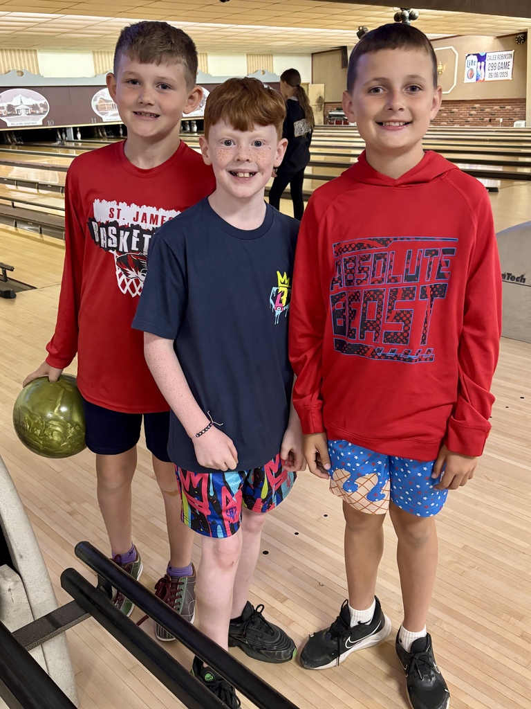 Three young boys stand at a bowling alley, smiling. Two wear red hoodies; one has a green bowling ball.
