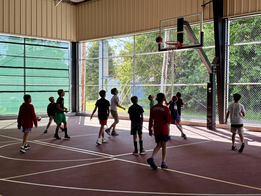 A group of children playing basketball in an indoor court with glass walls, allowing views of the outdoors.