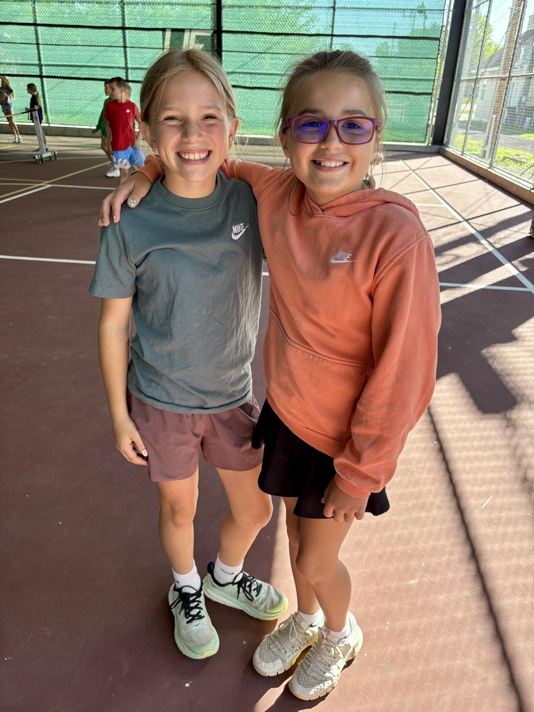 Two children stand close together on a tennis court, one with a gray t-shirt, the other in an orange hoodie.