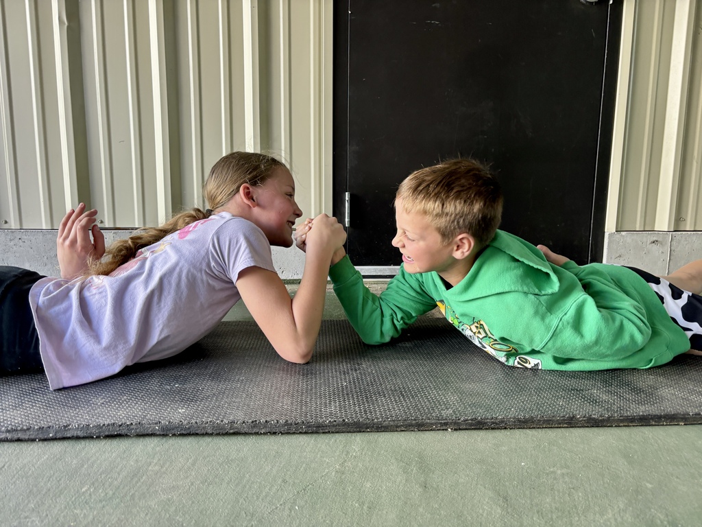 Two children arm wrestle while lying on the floor, a door behind them. The girl wears a white T-shirt, the boy a green hoodie.