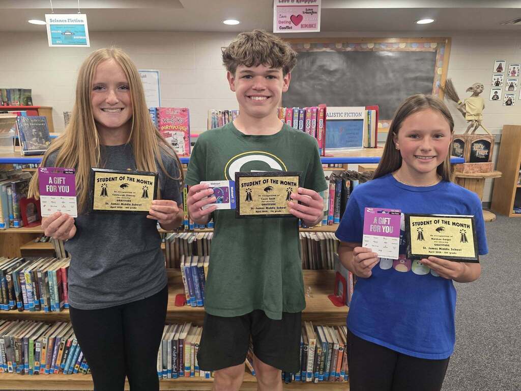 Three students, two girls and a boy, hold certificates in a library with bookshelves in the background.