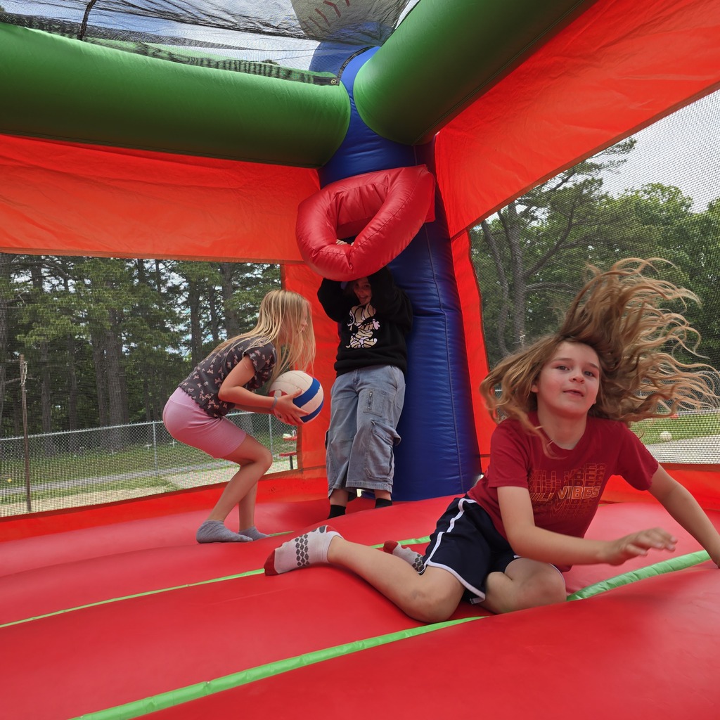 Three children play in a bouncy castle with a red floor, while trees and a fence are in the background.