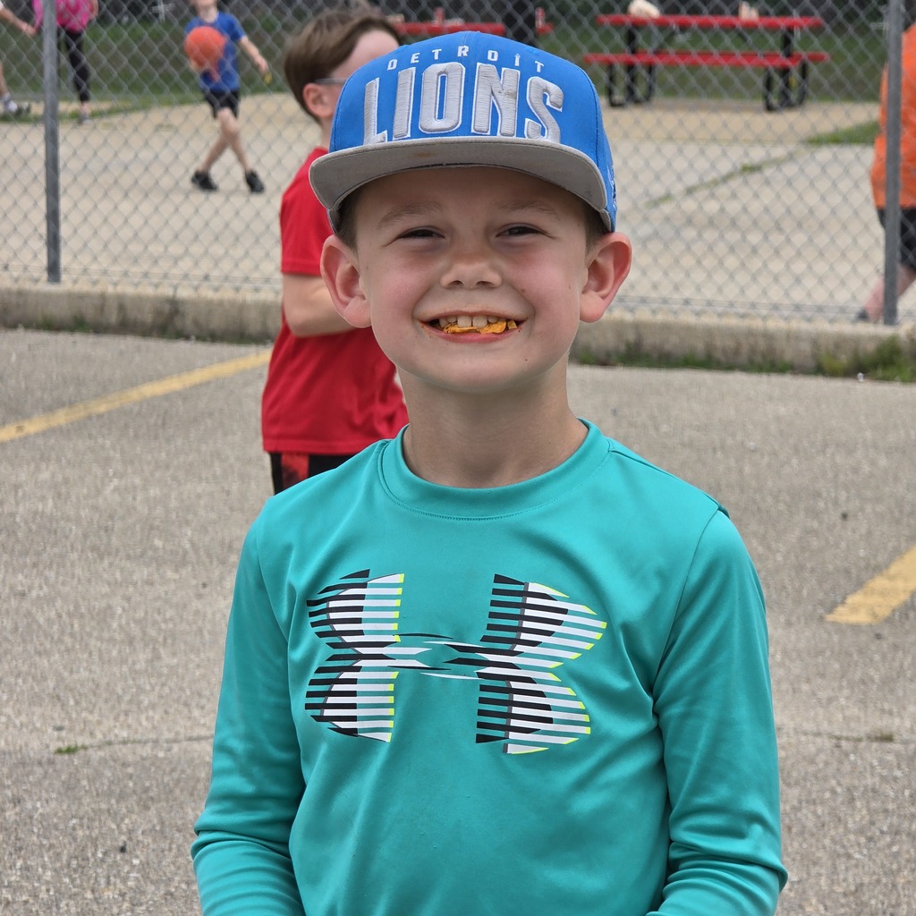 A boy in a teal long-sleeve shirt and a blue baseball cap stands in a park. People are walking and sitting nearby.