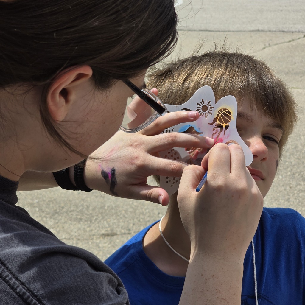A person applying face paint to a child with a design on paper. The child is wearing a blue shirt.
