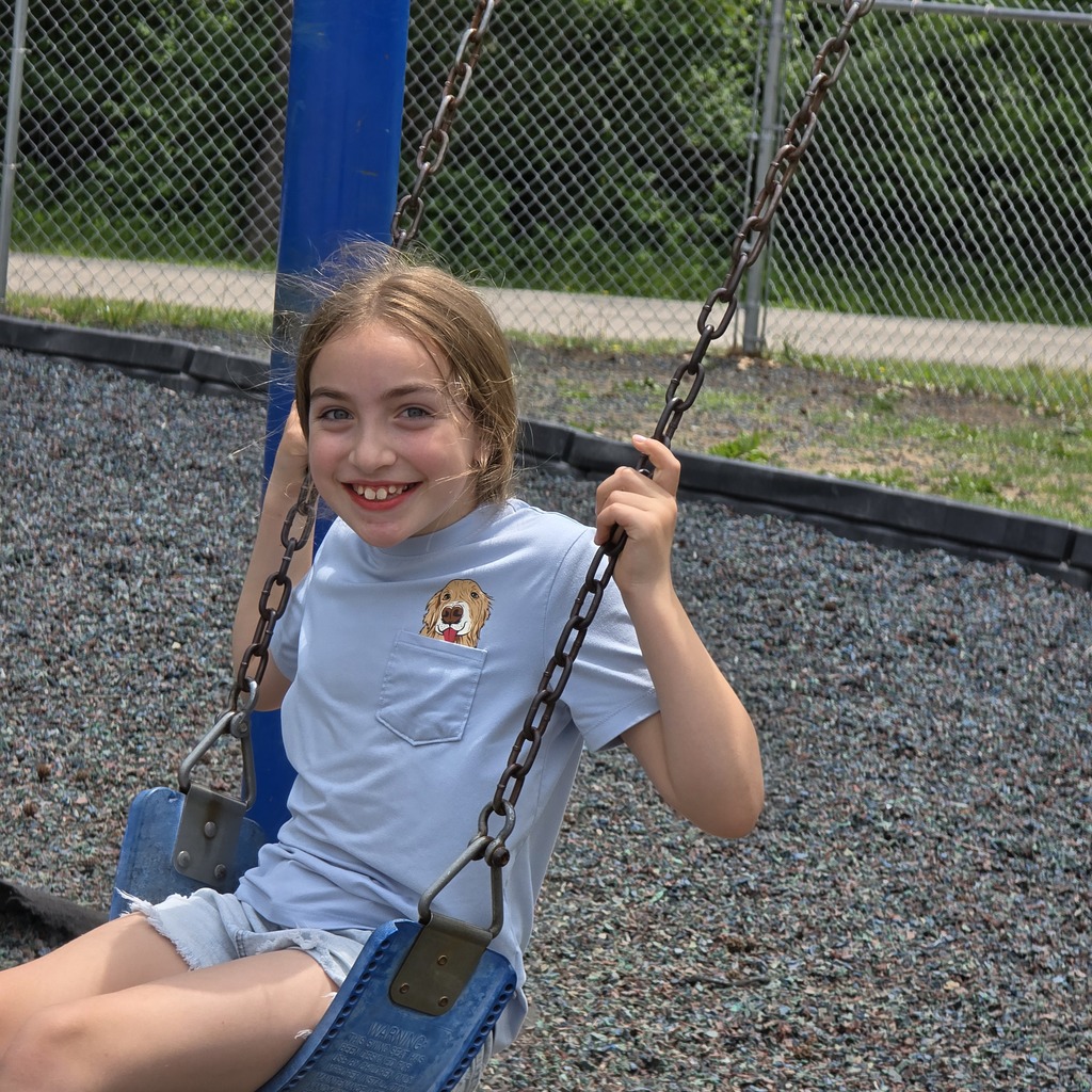 A girl with blonde hair sits on a blue swing in a park with gravel ground.