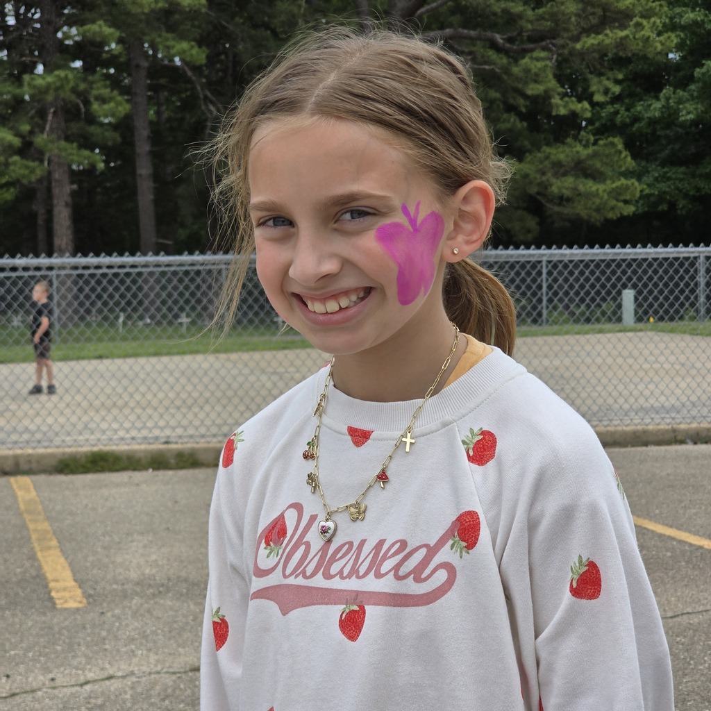 A young girl with a butterfly face painting smiles in a parking lot, wearing a strawberry-printed shirt.