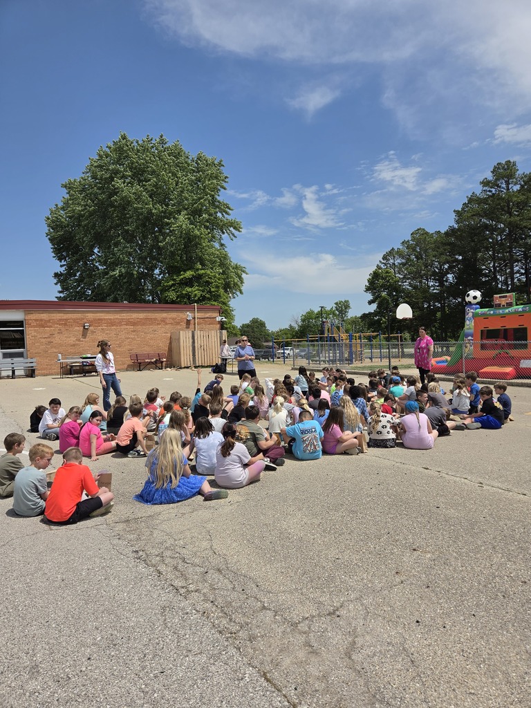 A group of children sits in a circle outdoors, with an adult standing in front. Trees and a building are in the background.