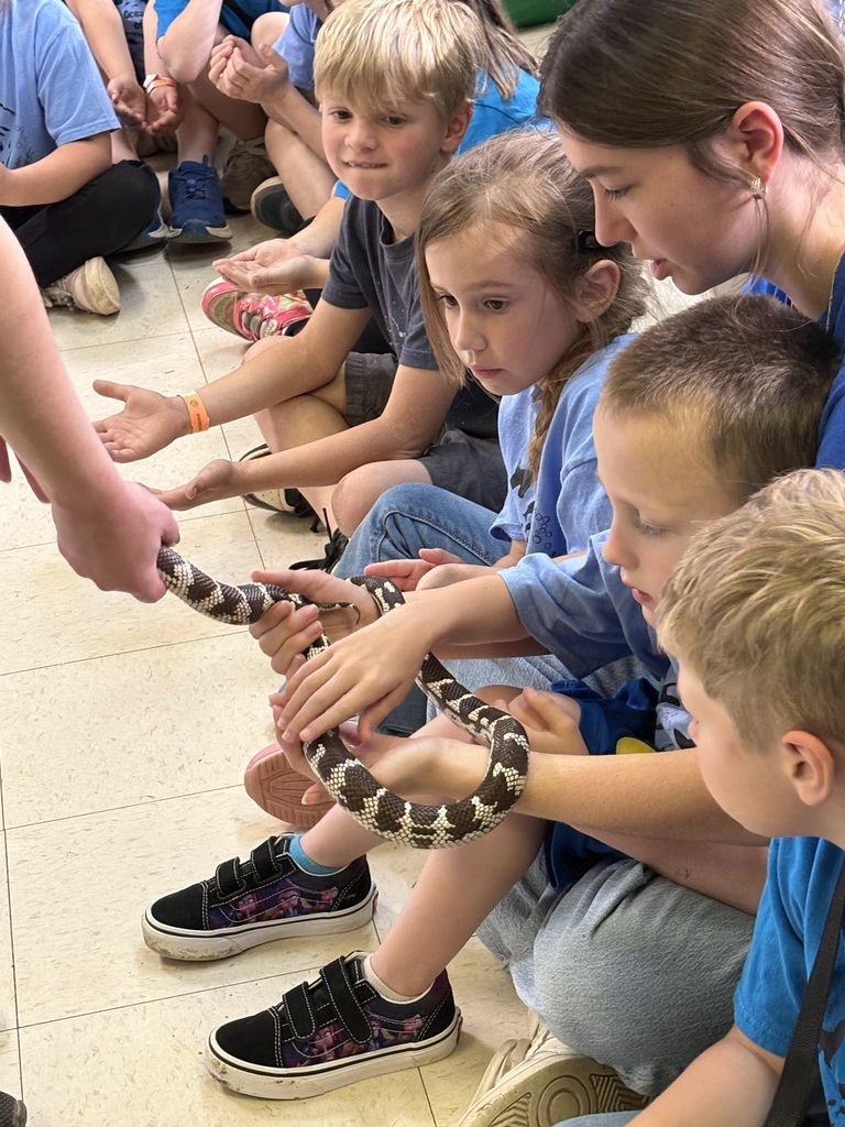 Several children, some wearing blue shirts, are seated around a woman who is holding a snake.