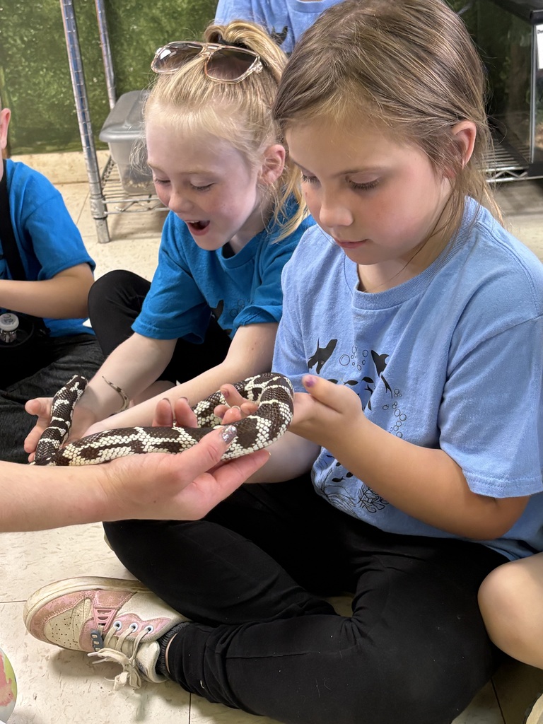 Kids sit on a floor, one holding a snake, others watching. They wear blue shirts and black pants.