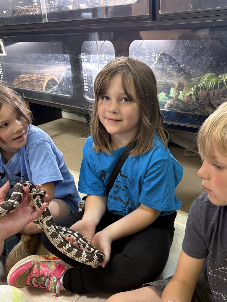 Three kids sit on the floor. A girl holds a snake. Behind them is a glass wall showing reptiles in cages.