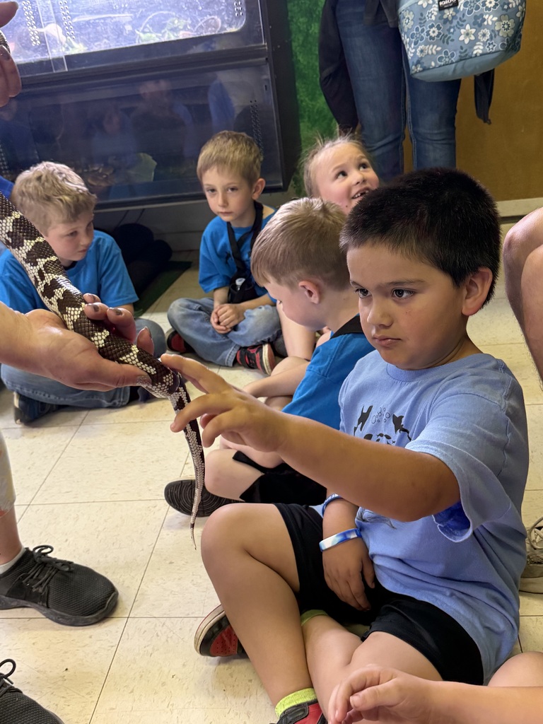 Children sit on floor in circle, holding snake as it slithers on hands. Light wood floor, glass window, white walls.