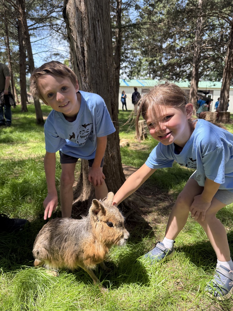 Two kids in blue shirts stand by a small animal. One holds the animal. Trees are in the background.